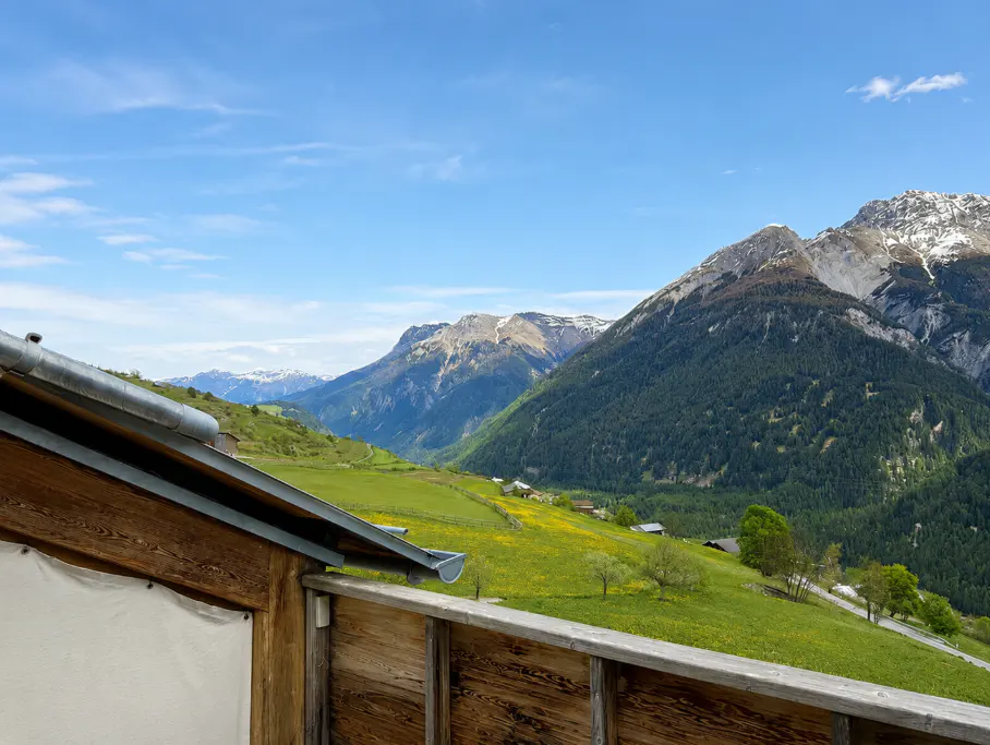 View from a wooden balcony overlooking a green valley and mountains under a blue sky.