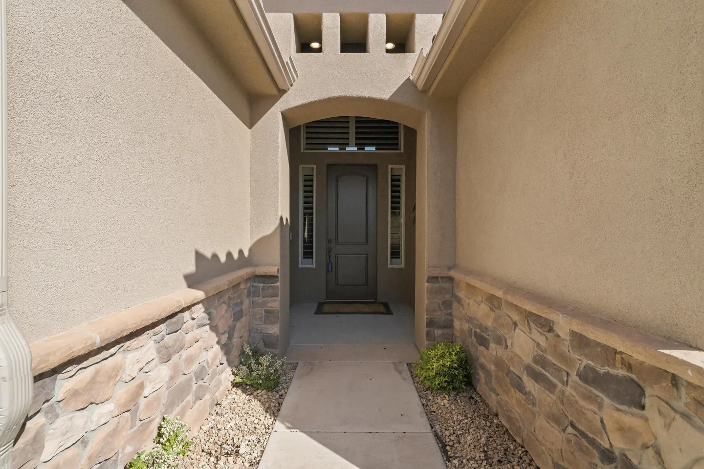 A concrete walkway leads to a dark brown front door framed by tan stucco walls and decorative stone accents.