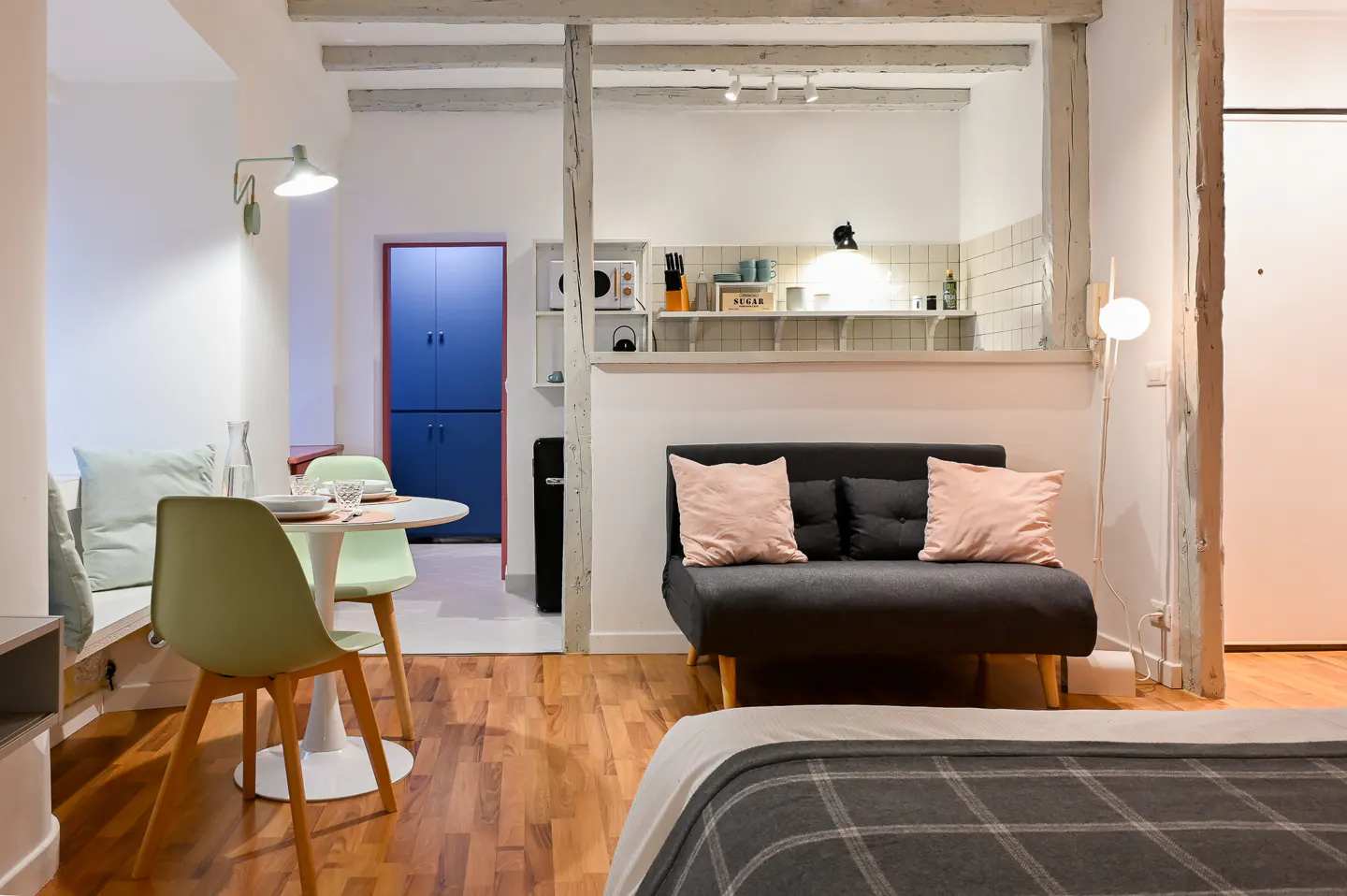 Studio apartment with hardwood floors, a gray sofa with pink pillows, and a dining area with green chairs. A small kitchen is visible in the background.