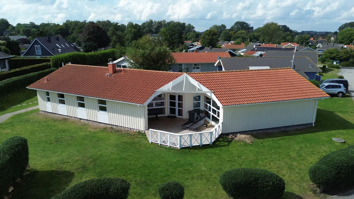 Aerial view of a one-story, white house with a red tile roof and a wooden deck, surrounded by green grass and trees.