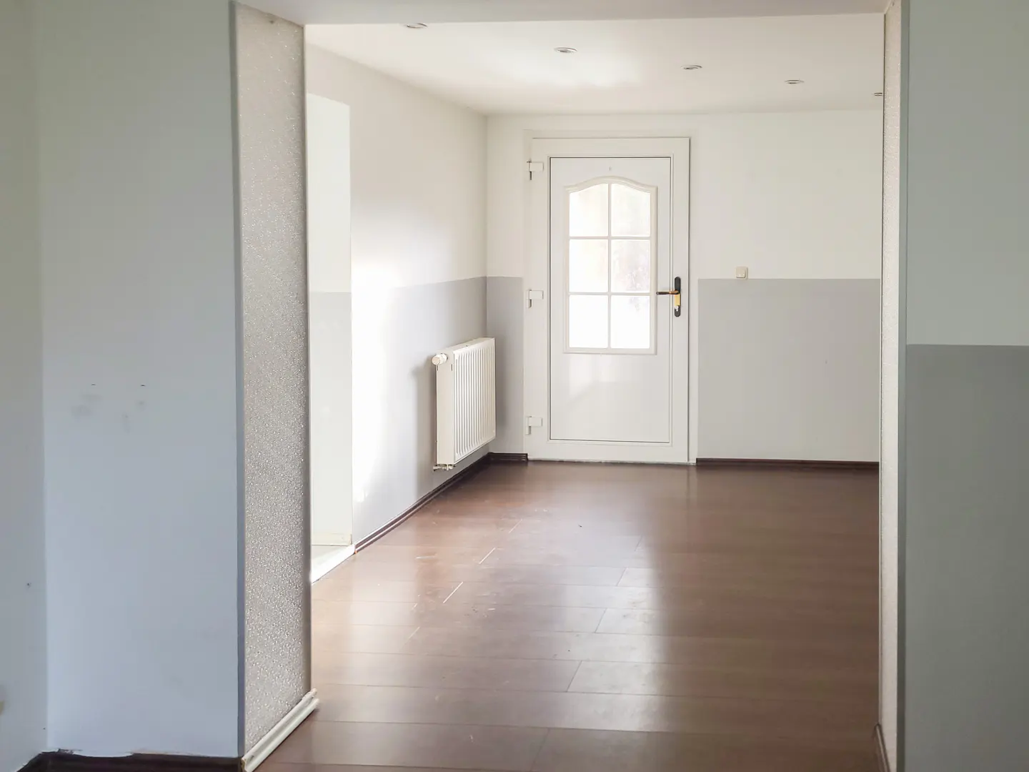 Interior view of a room with white walls, a radiator, and a white door with glass panels. The floor is brown wood.