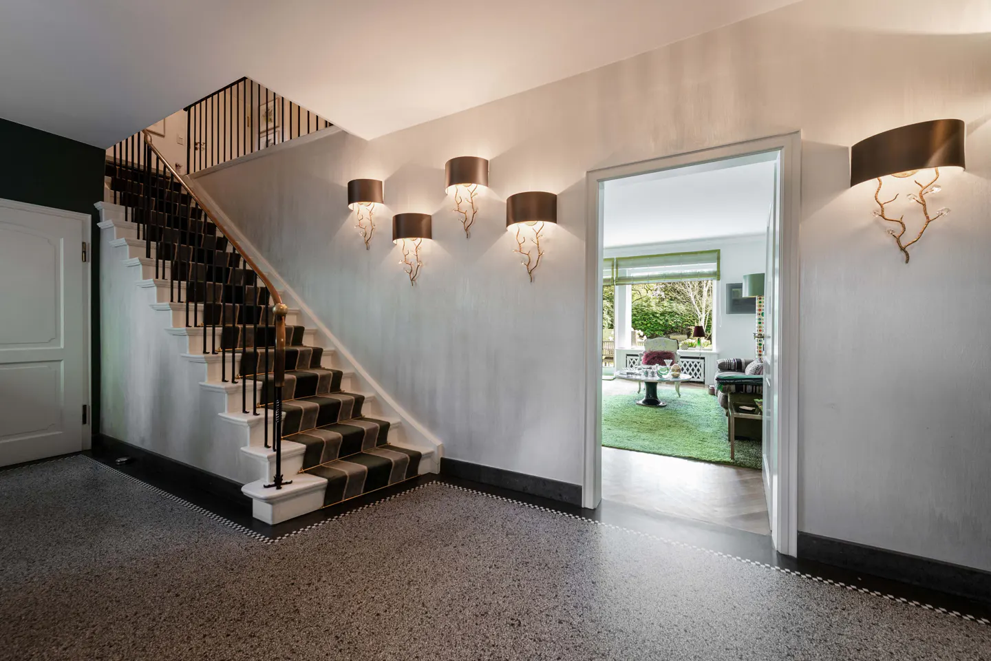 Entryway with a striped staircase, speckled flooring, and decorative wall sconces, leading to a bright living room.