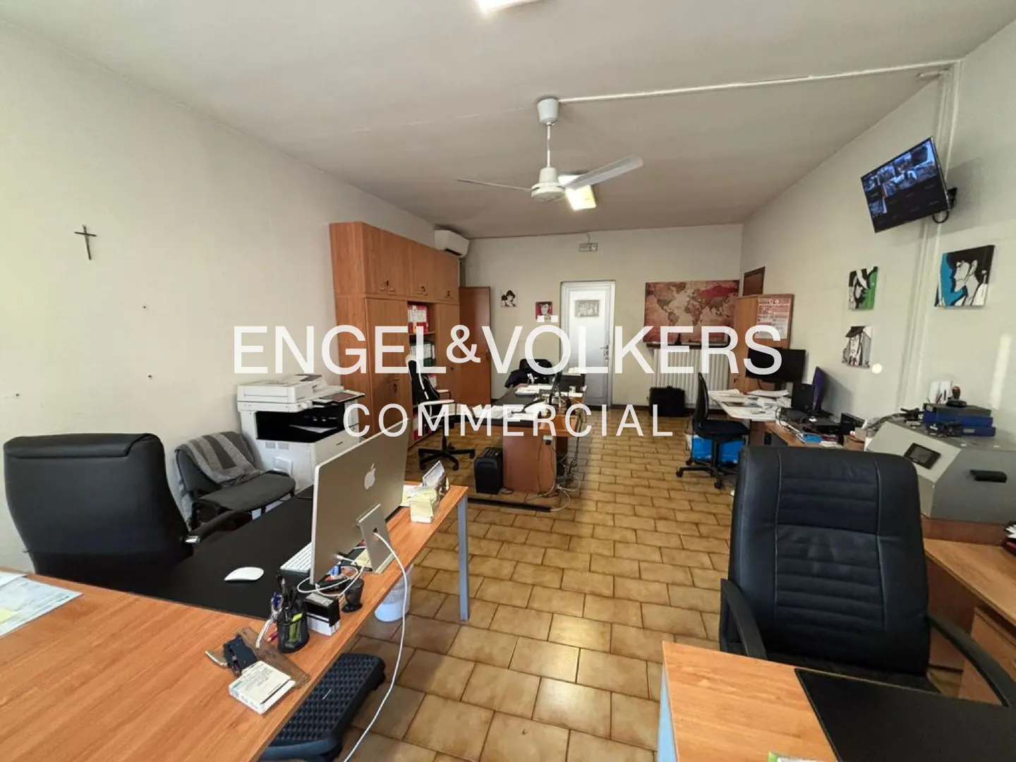 A wide shot of a commercial office space with desks, chairs, and computers. The walls are white, and the floor is tiled.