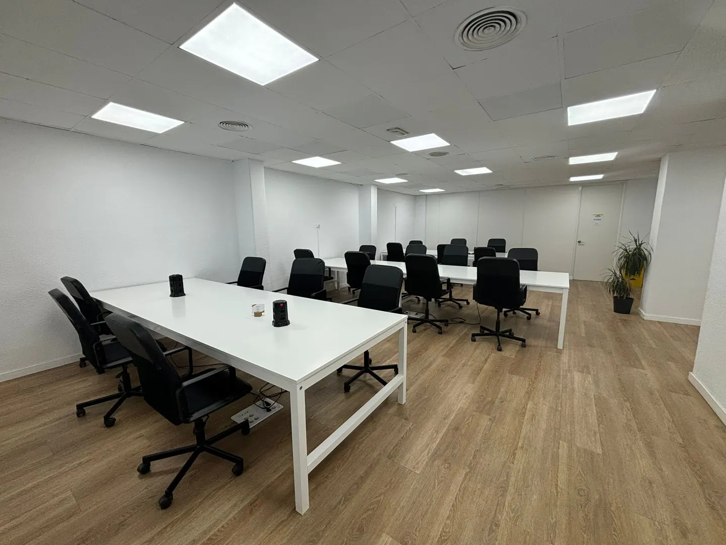 Bright office meeting room with white tables, black rolling chairs, wood-look flooring, and recessed ceiling lights.