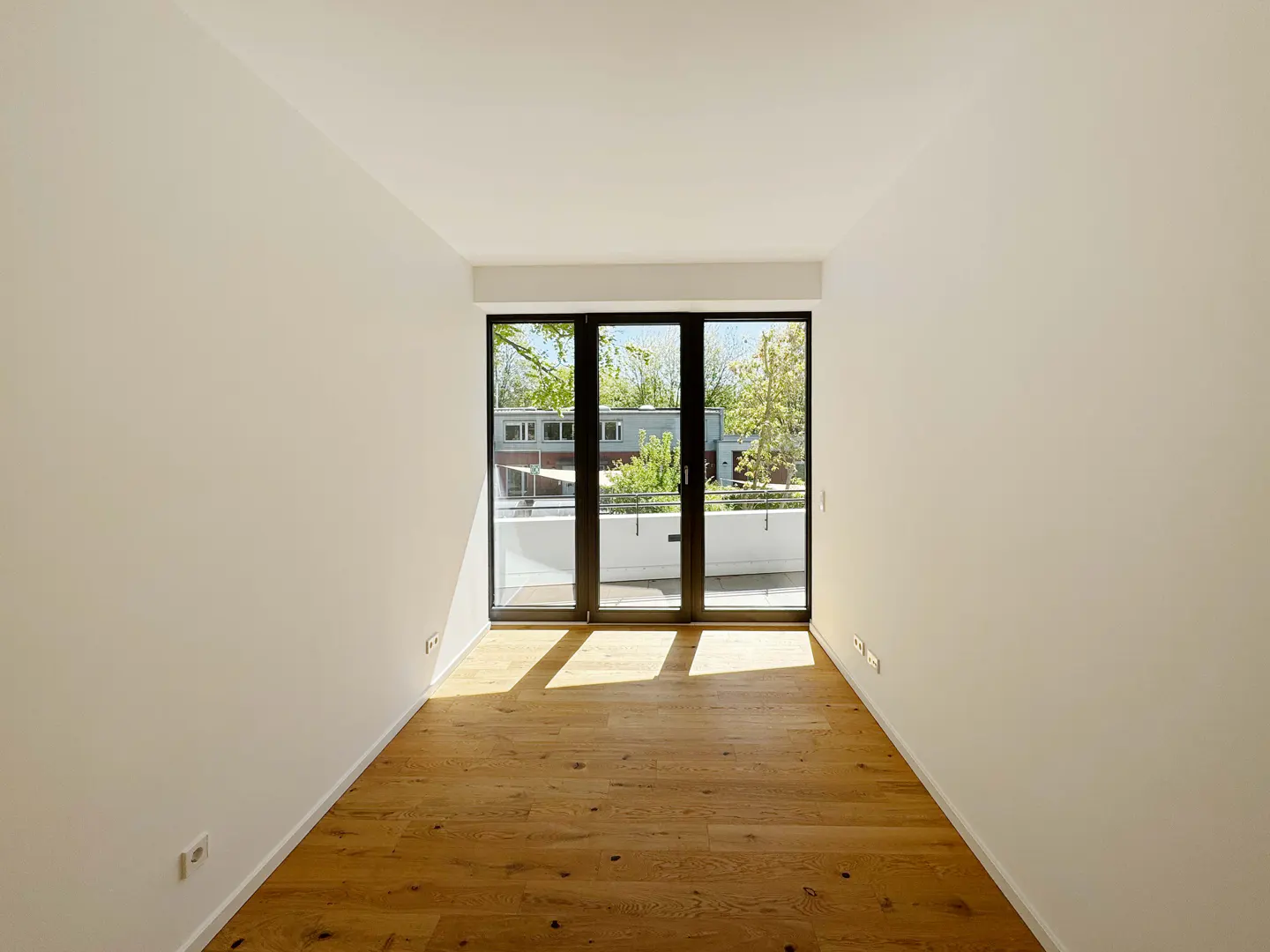 Empty room with white walls, light wood flooring, and large black-framed glass doors leading to a sunny balcony.