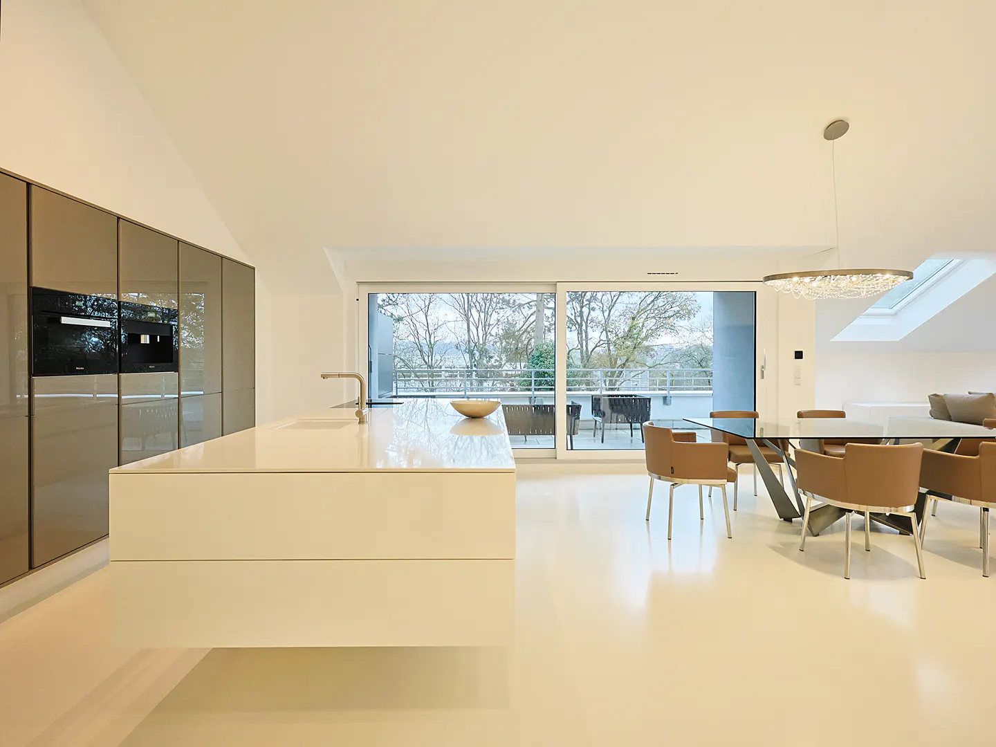 Bright, modern kitchen and dining area with white floors, island, and glass table. Balcony view with trees.