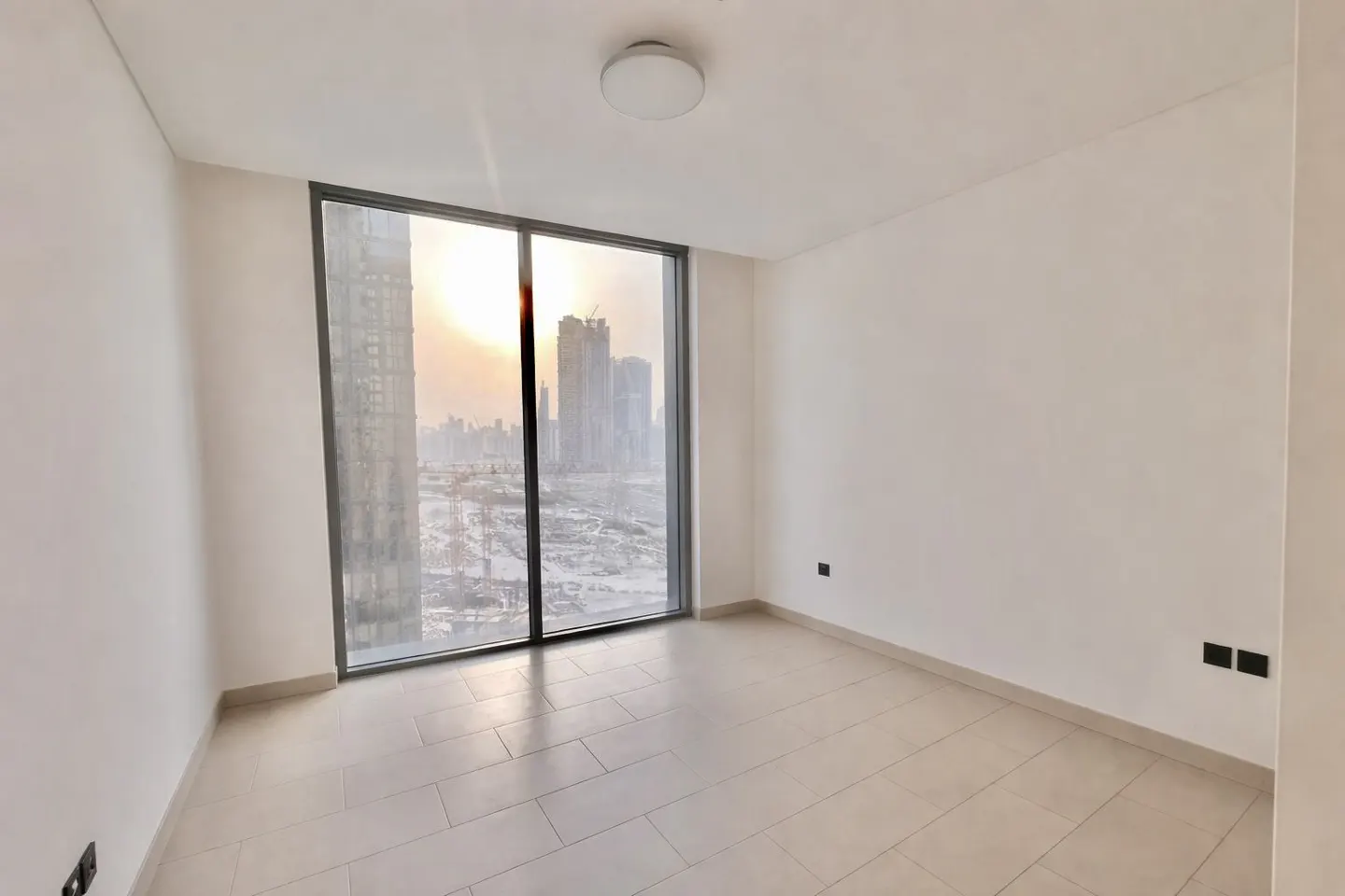 Empty room with white walls, light tile floor, and large window showing a city skyline at sunset.