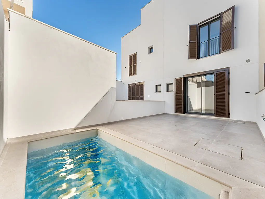 A modern white house with a small pool and a gray patio under a blue sky. Brown shutters adorn the windows.