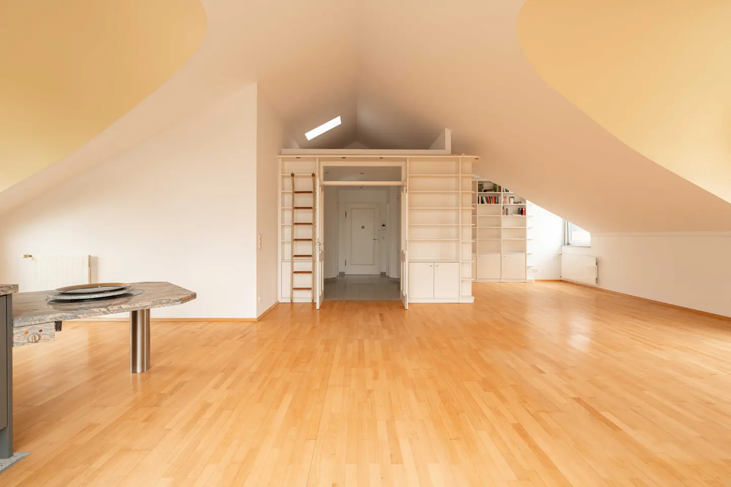 Attic apartment with light wood floors, white walls, and a yellow ceiling. A white bookcase with a ladder surrounds a doorway.