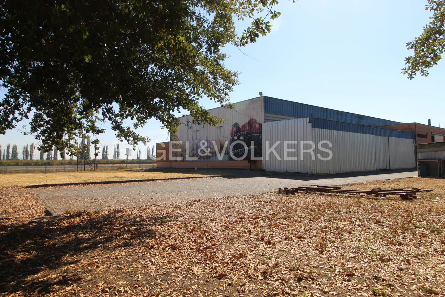 Exterior view of a commercial building with Engel & Volkers logo, a blue roof, and a gravel lot covered in leaves.