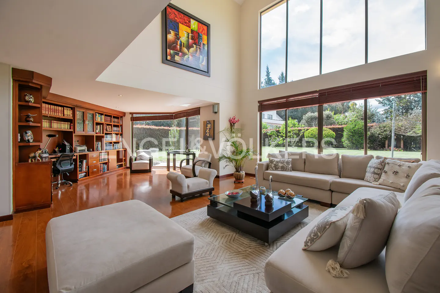 Bright living room with beige sofas, a black coffee table, and a wooden bookcase. Large windows overlook a green garden. An abstract painting hangs on the wall.