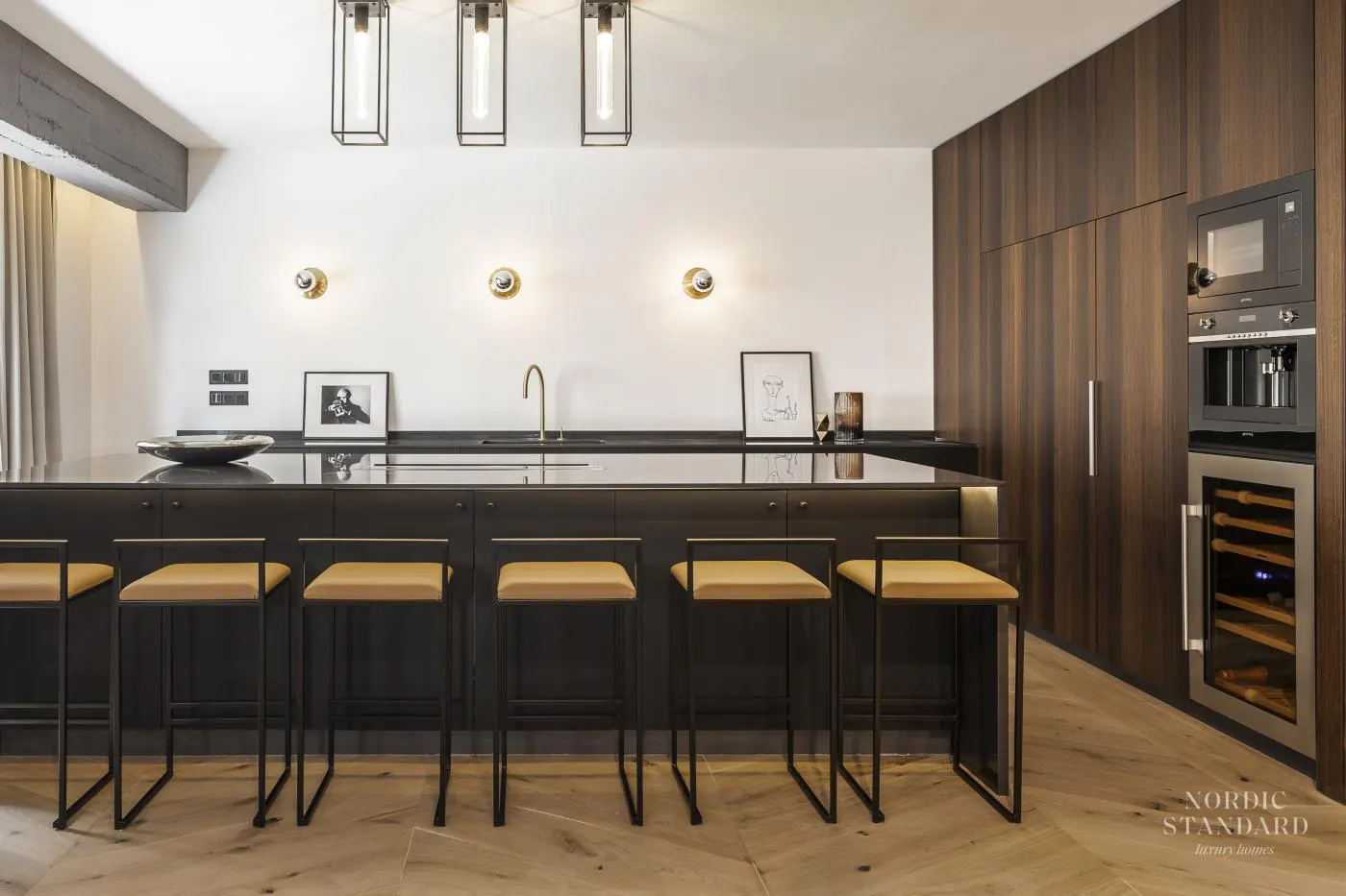 A modern kitchen with black island, tan stools, wood cabinets, and stainless steel appliances. Three pendant lights hang above.