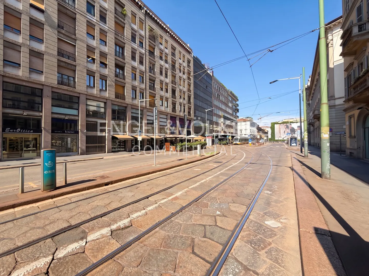 A sunny city street with tram tracks running along a stone-paved road, flanked by tall historic buildings and shops.