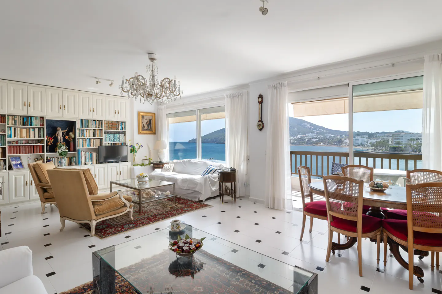 Bright living room with white walls, a chandelier, and a view of the ocean. There are chairs, a sofa, and a dining table.