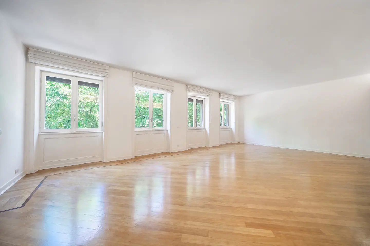 Bright, empty room with light wood floors, white walls, and four large windows overlooking green trees.