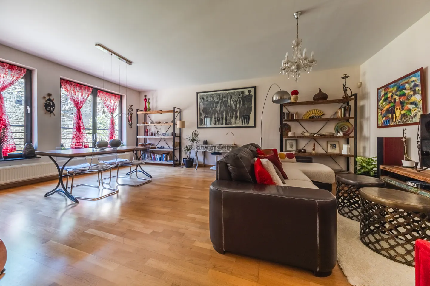 A bright living room with hardwood floors, a brown leather sofa, and a dining table with red curtains. Shelves display books and decor.