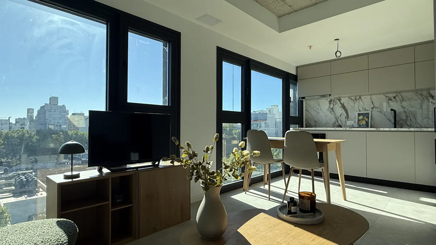 Bright apartment interior with black-framed windows, a TV, a dining table with two chairs, and a modern kitchen with marble backsplash.
