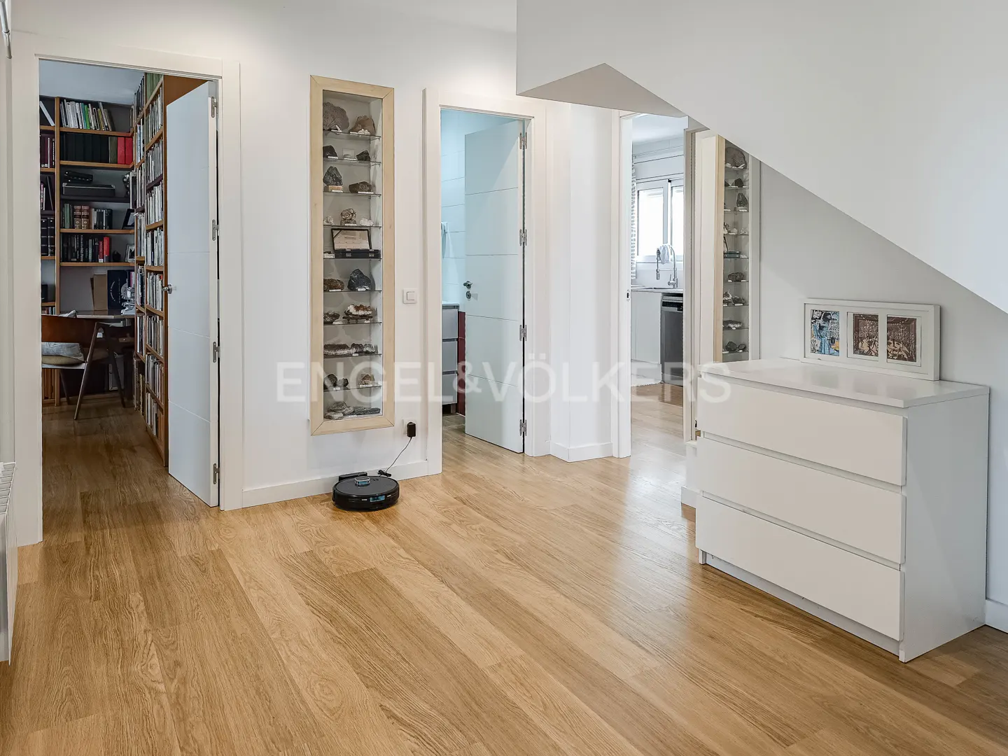 Bright hallway with light wood floors, white walls, a white dresser, a wall-mounted display case, and a robot vacuum.