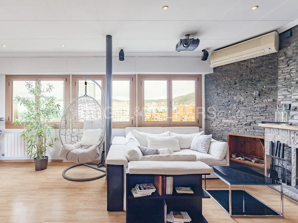 Bright living room with wood floors, stone wall, and large windows. A white sofa, hanging chair, and black shelving unit furnish the space.