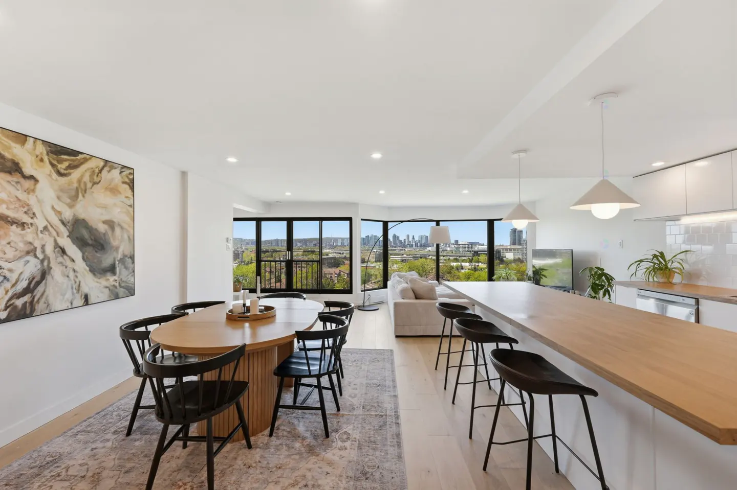 Bright, open-concept living space with dining table, kitchen island with stools, and city views through large windows. Neutral color palette.