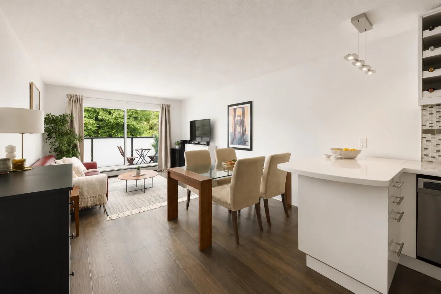 Bright, open-concept living space with wood floors, dining table, and balcony with green view. Modern kitchen island visible.