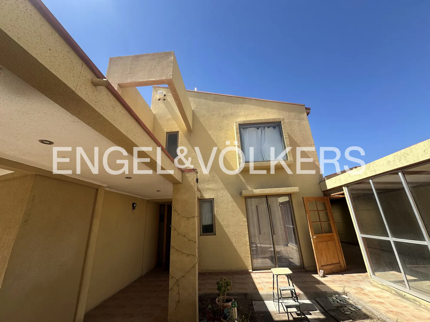 Exterior view of a two-story tan house with an open door, a small stool, and a sunroom under a clear blue sky.