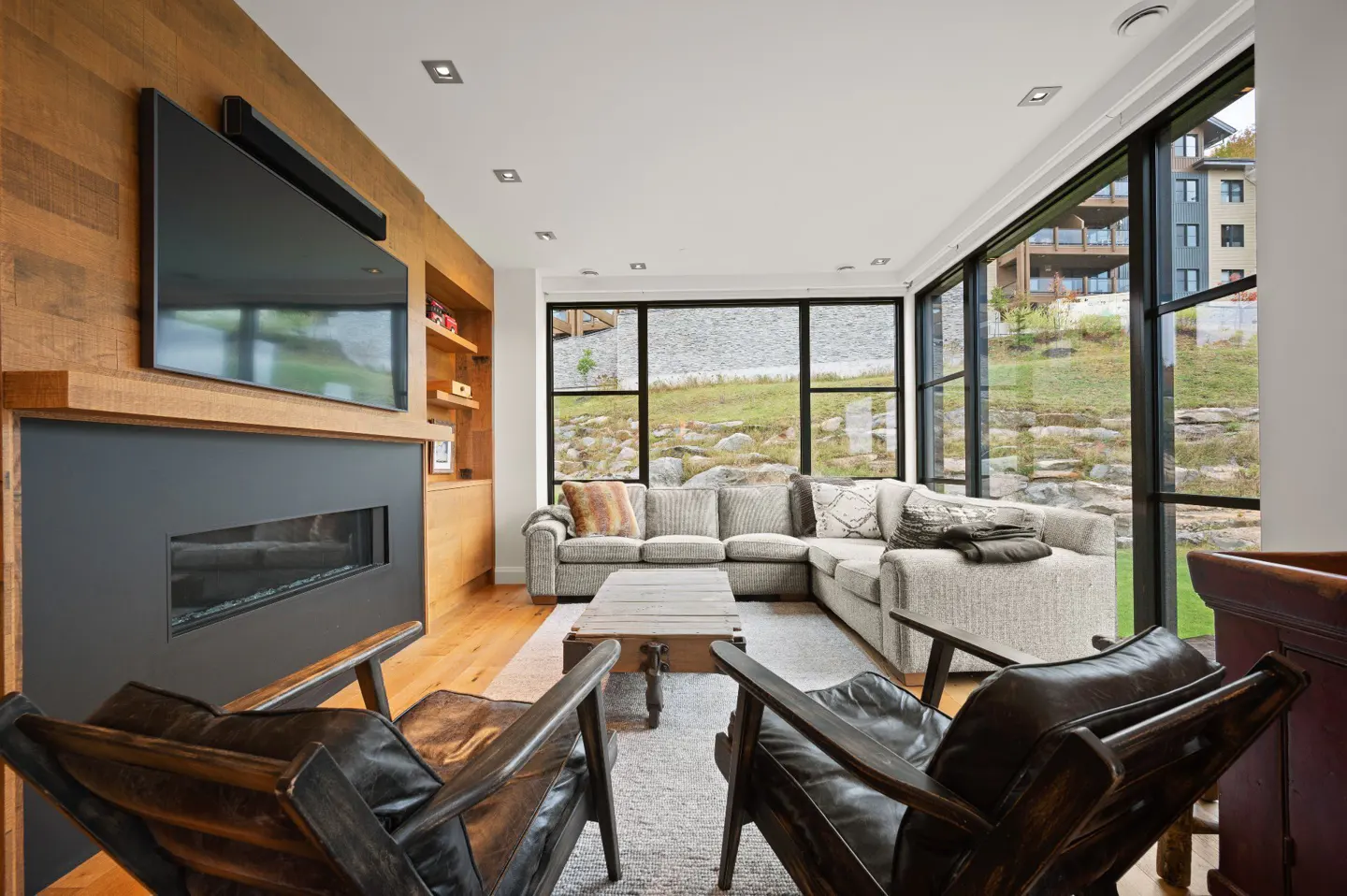 Living room with a gray sectional sofa, two leather chairs, a fireplace, and a large window with a view of a lake.