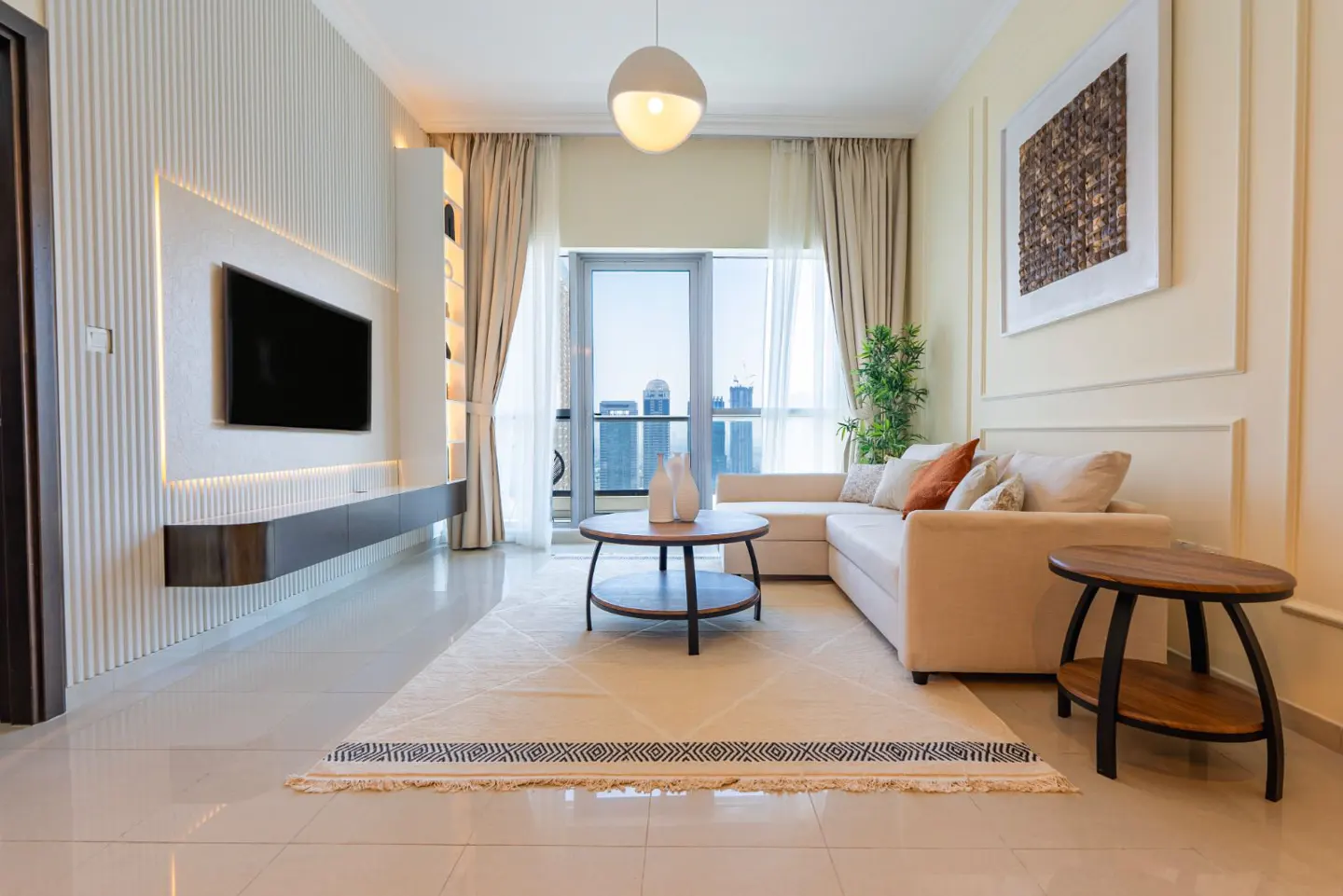 Bright living room with a cream L-shaped sofa, round wood tables, and a large TV mounted on a textured wall. A rug sits on the tiled floor.