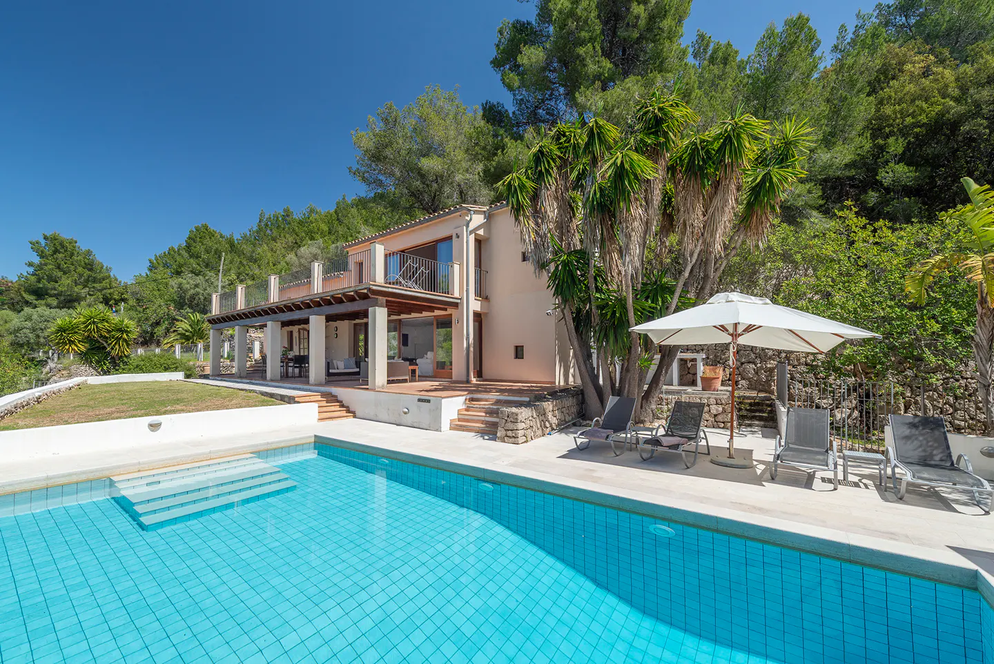 Exterior of a two-story beige house with a blue tiled pool, lounge chairs, and green trees under a clear blue sky.