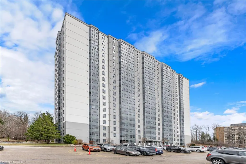 Exterior view of a tall, gray and white apartment building with many windows, a parking lot, and a blue sky.
