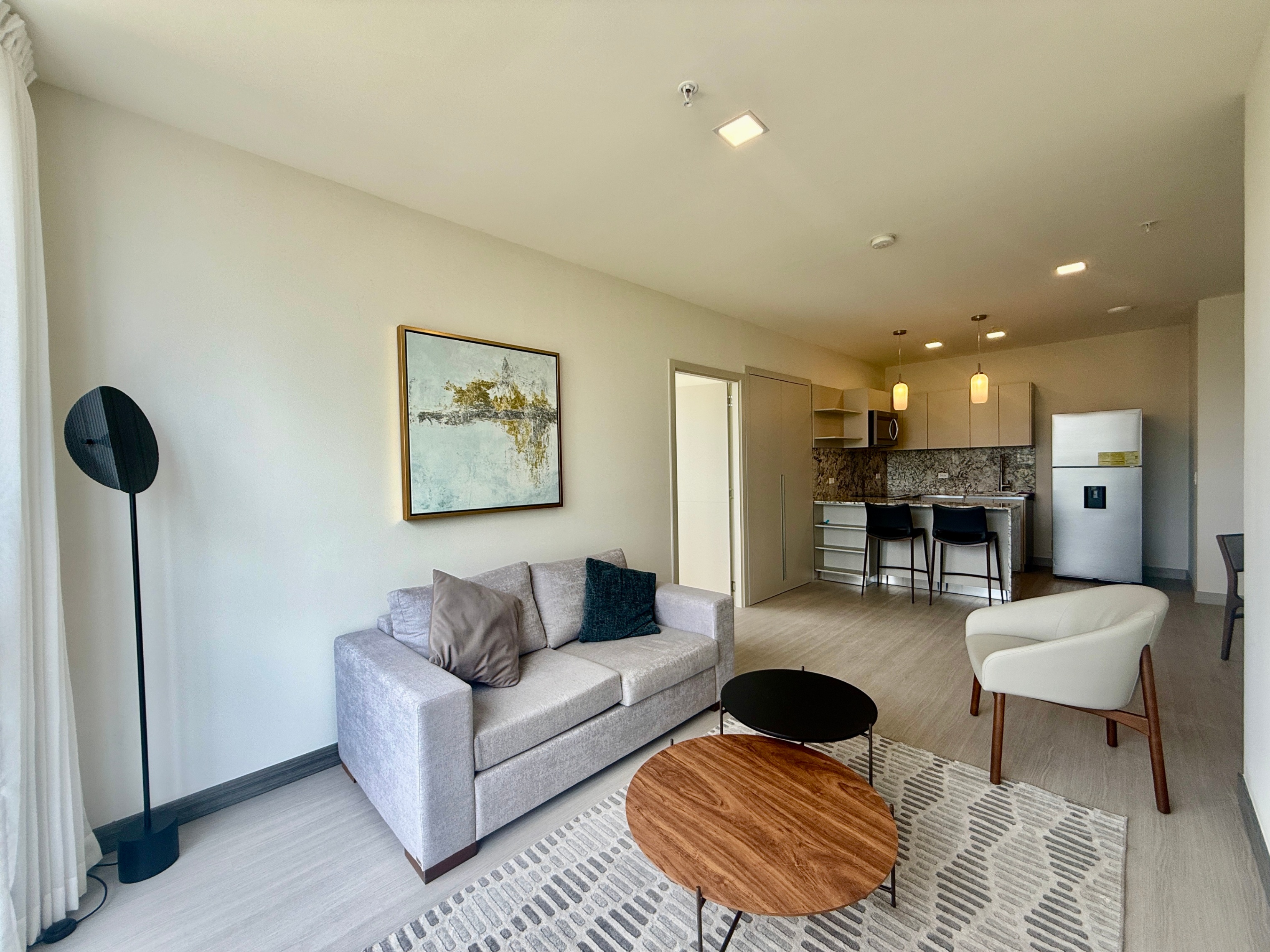 Living room with gray sofa, wood tables, and abstract art. Kitchen with island and stools in the background.
