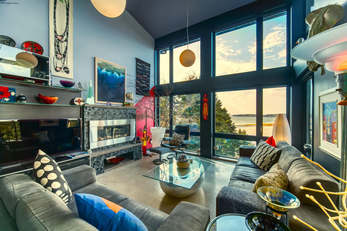 Modern living room with a fireplace, glass coffee table, grey sofas, and floor-to-ceiling windows overlooking the ocean.