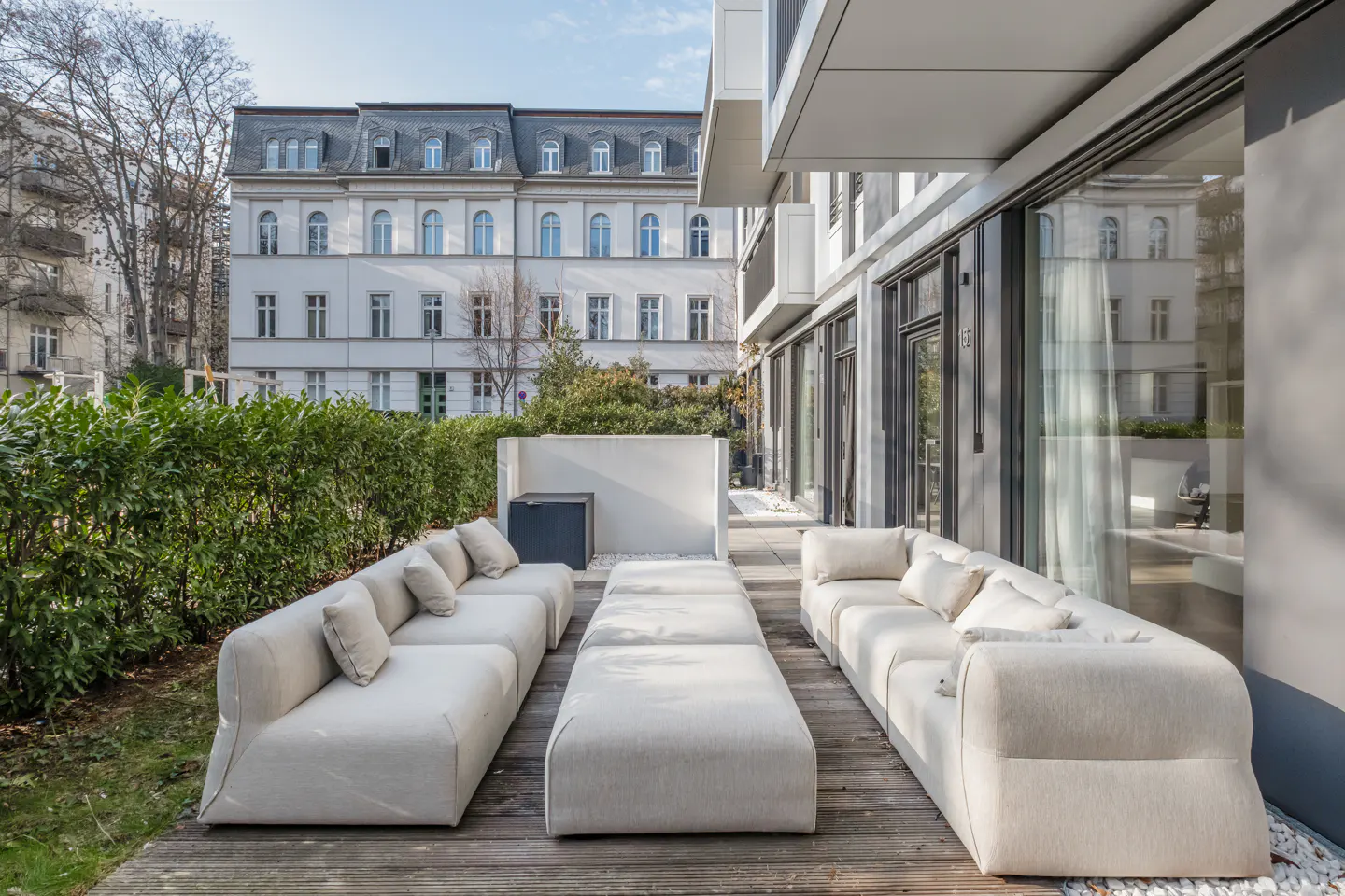 Outdoor patio with two cream sofas, ottomans, and green hedges. A white building is in the background.