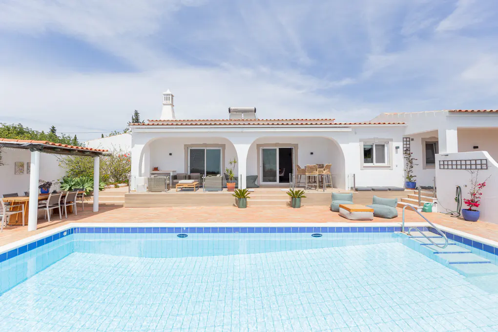 A white villa with a red tile roof overlooks a blue tiled pool on a sunny day. Outdoor seating is visible.