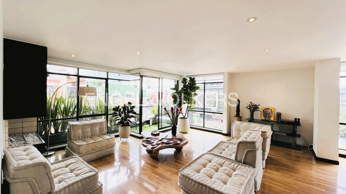 Bright living room with wood floors, white sofas, and large windows overlooking greenery. A unique wood coffee table sits in the center.
