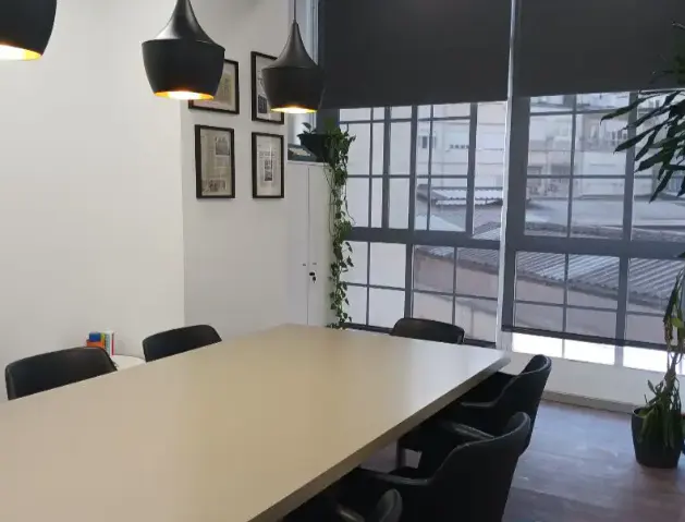 Conference room with a long table, black chairs, and black pendant lights. Large windows with gray shades and framed pictures on the white wall.