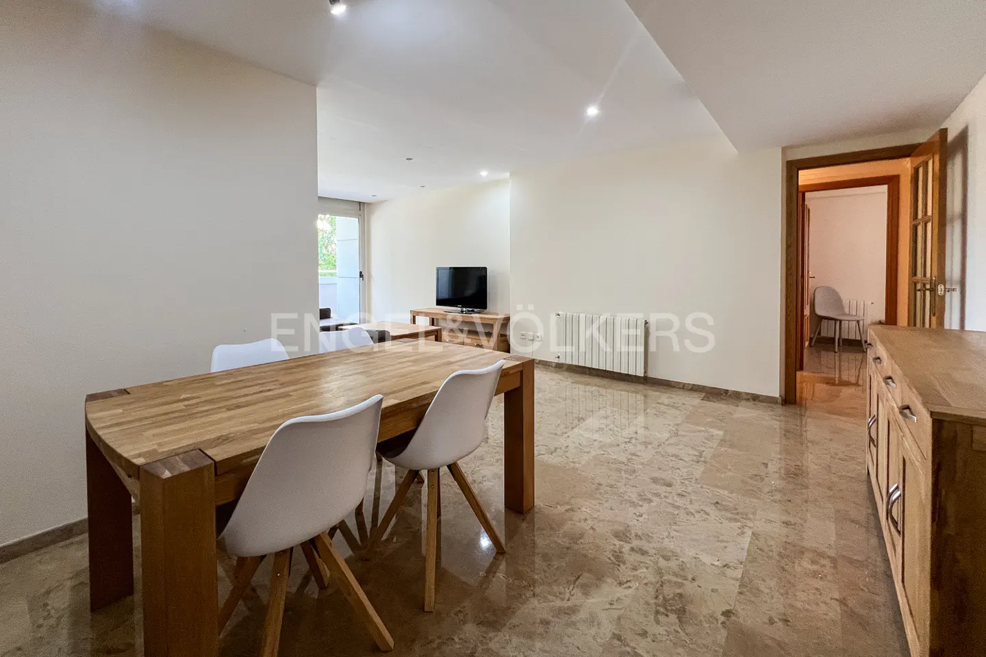 Open-concept living and dining area with marble floors, wooden table and white chairs. A TV and cabinet are in the background.