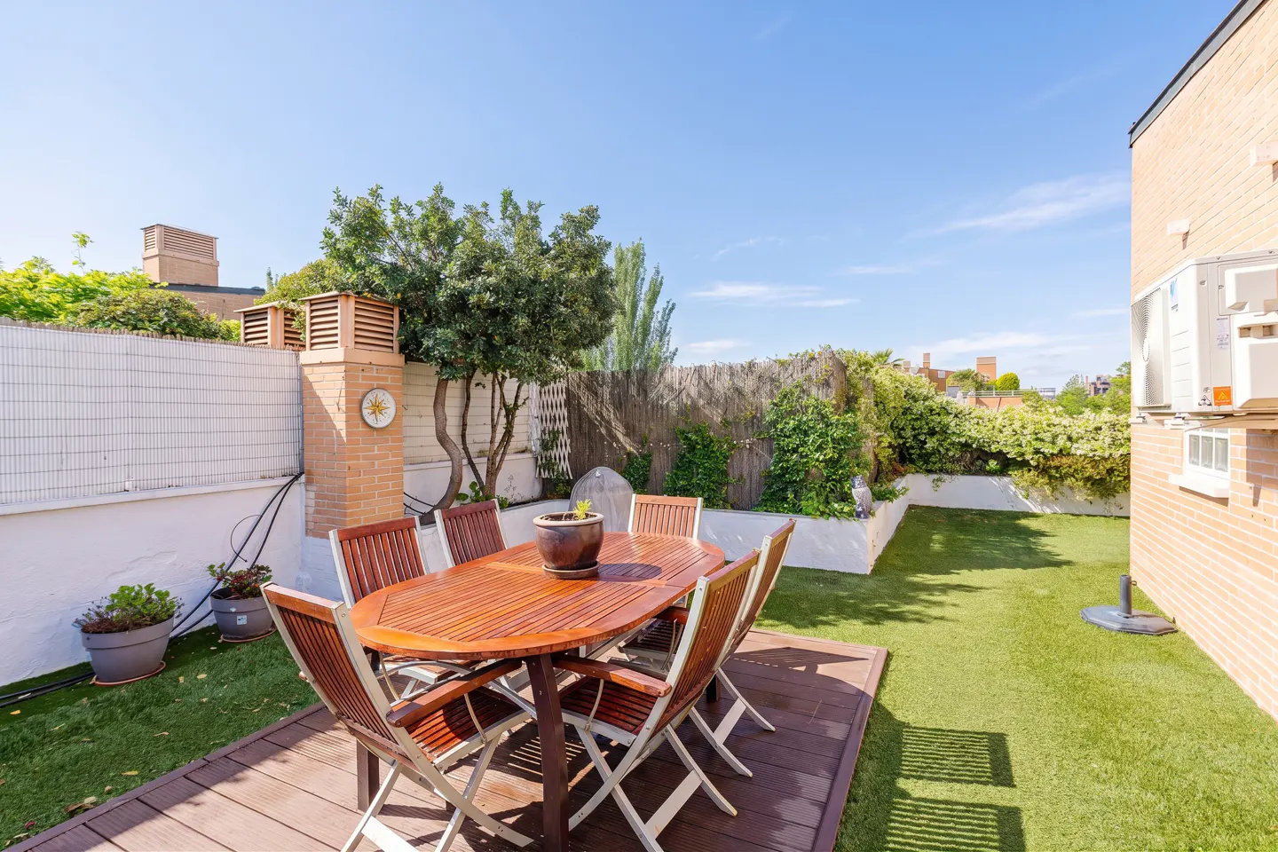 Outdoor patio with a wooden table and chairs on a deck, surrounded by green grass and plants.