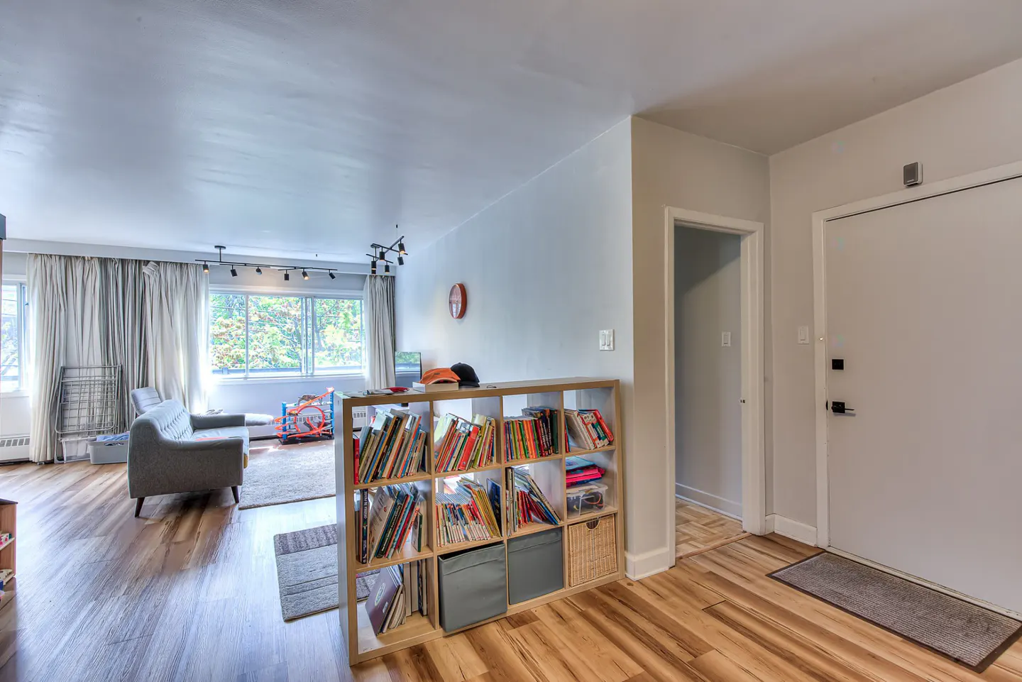 A bright living room with wood floors, a gray sofa, and a bookshelf filled with books and storage bins. A white front door is visible.
