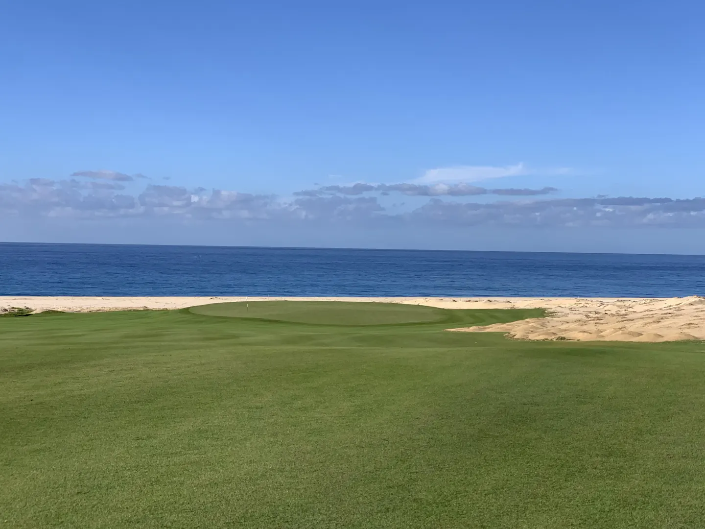 A green golf course fairway leading to a putting green, bordered by sand dunes and the blue ocean under a clear sky.