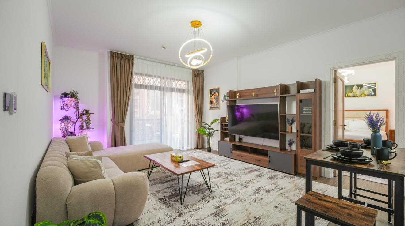 Living room with beige sectional sofa, wood coffee table, TV, and dining table set for four. A bedroom is visible through an open doorway.