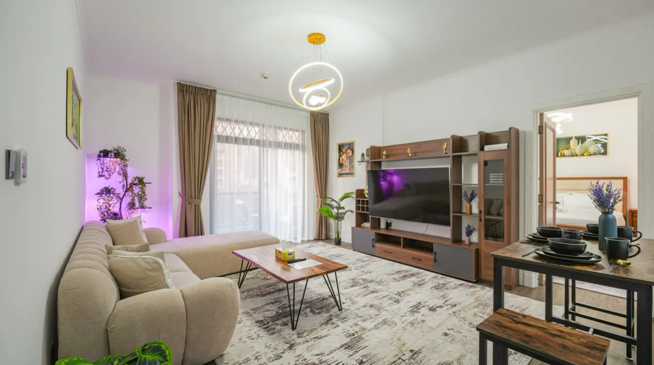 Living room with beige sectional sofa, wood coffee table, TV, and dining table set for four. A bedroom is visible through an open doorway.
