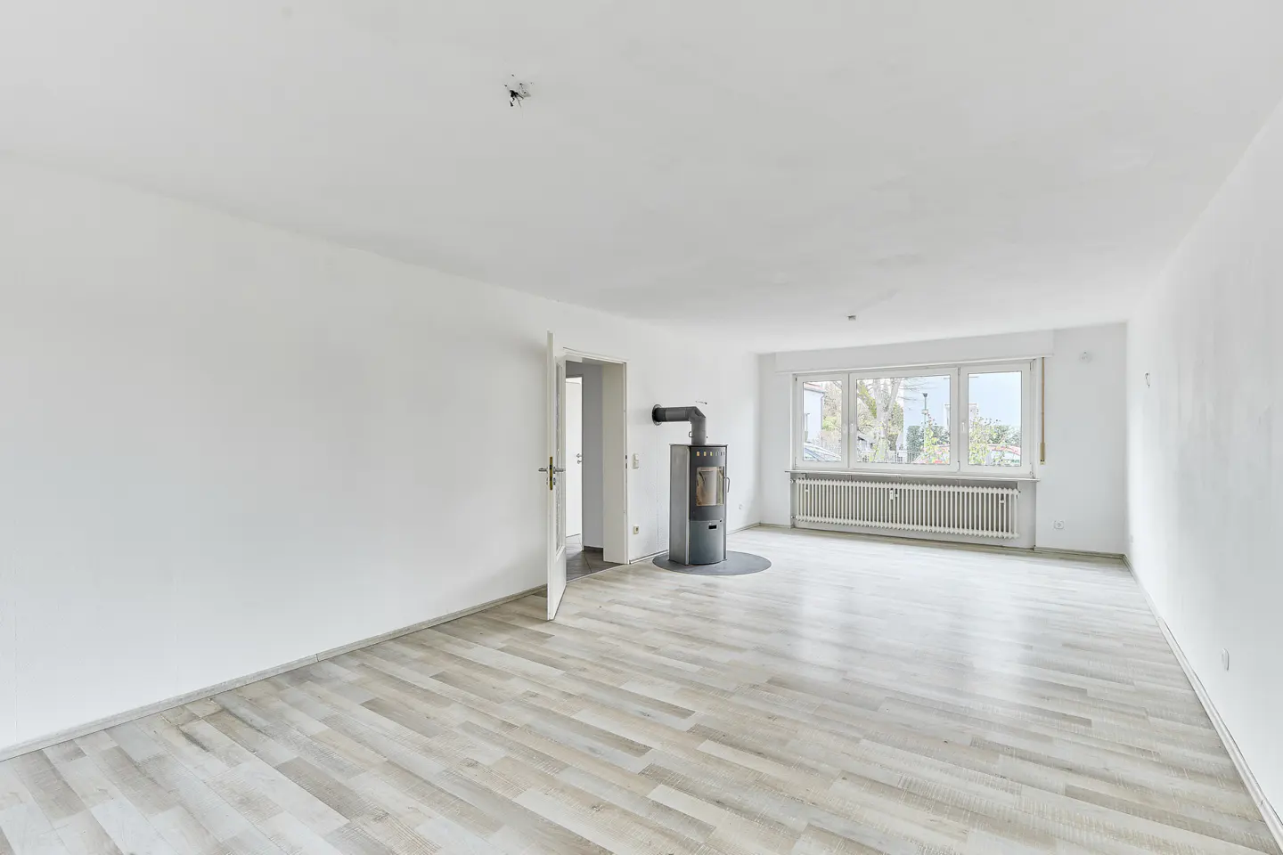 Bright, empty room with light wood floors, white walls, and a gray wood-burning stove. A window and open door let in natural light.