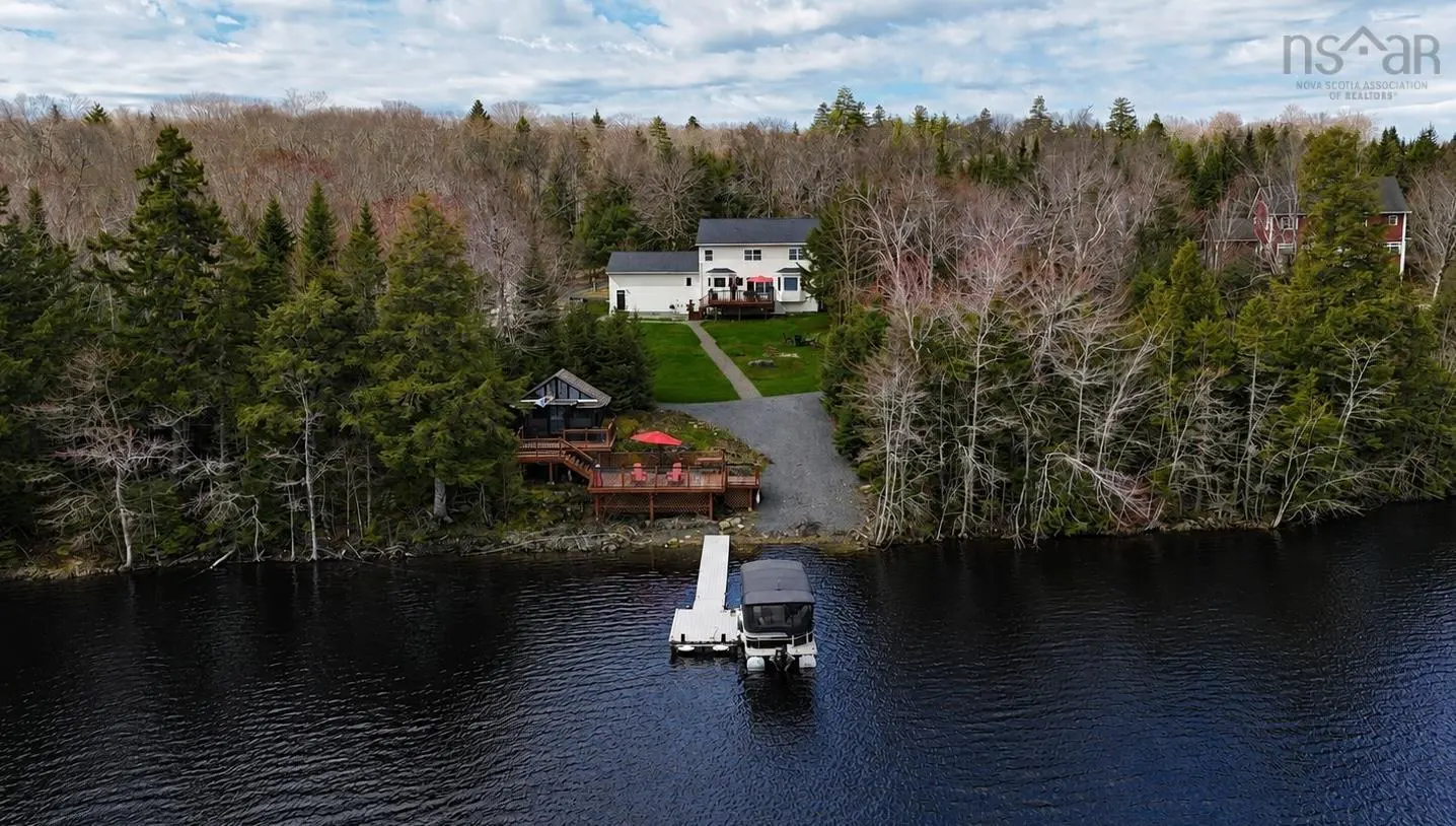 Aerial view of a white house and a cabin on a lake with a boat docked at a pier. The property is surrounded by trees.