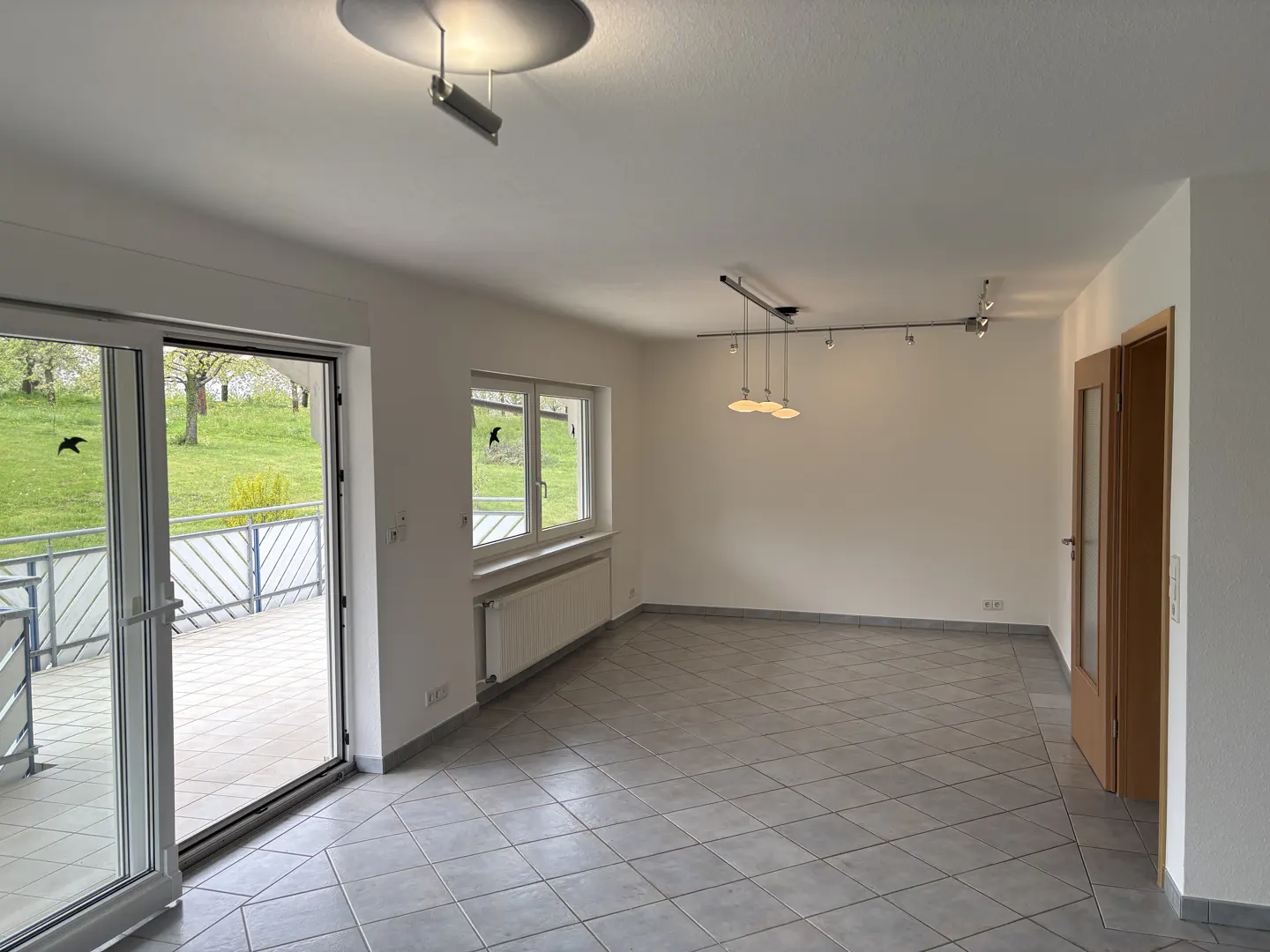 Bright, empty room with gray tile floor, white walls, and a sliding glass door to a balcony with a green view.