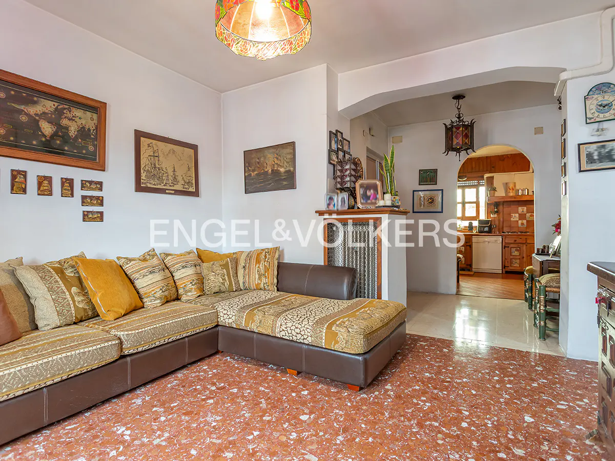 Living room with brown leather sectional sofa, patterned cushions, and red terrazzo floor. Archway leads to kitchen. Artwork on white walls.