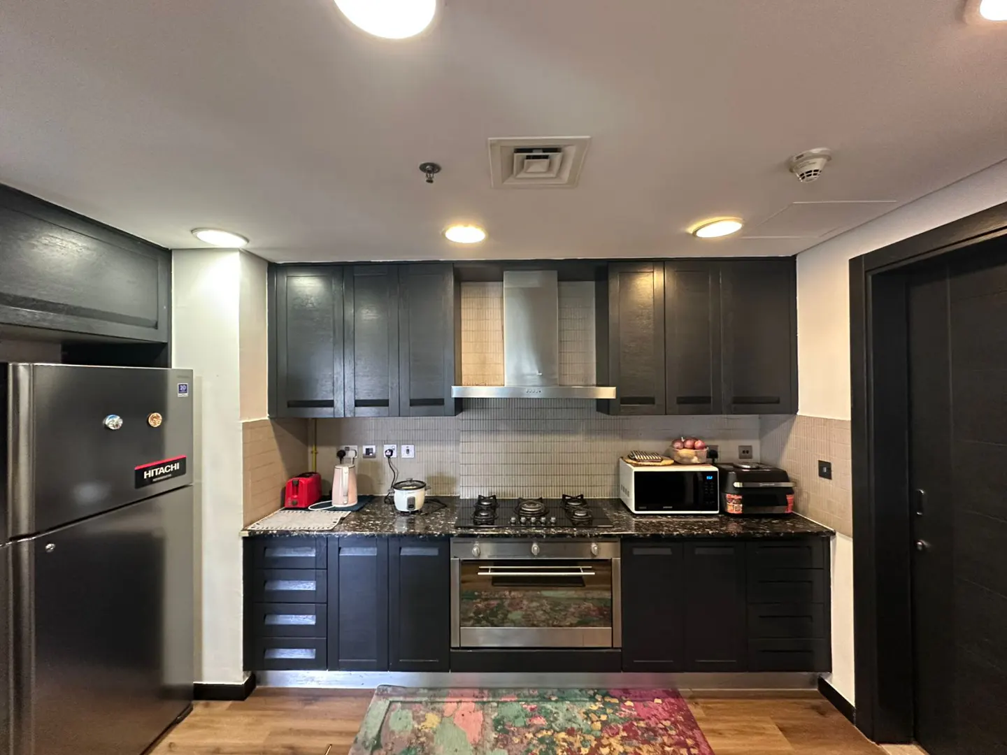 A kitchen with dark cabinets, stainless steel appliances, and a colorful rug on the wooden floor.