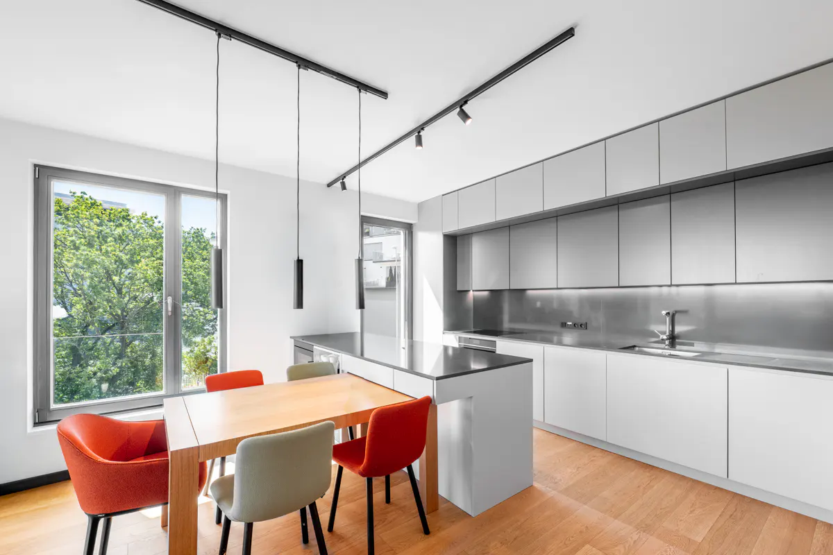 Modern kitchen with white cabinets, stainless steel backsplash, wooden dining table, and colorful orange and grey chairs.