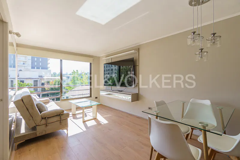 Bright living room with a beige sofa, glass coffee table, TV, and glass dining table with white chairs. Large window with city view.