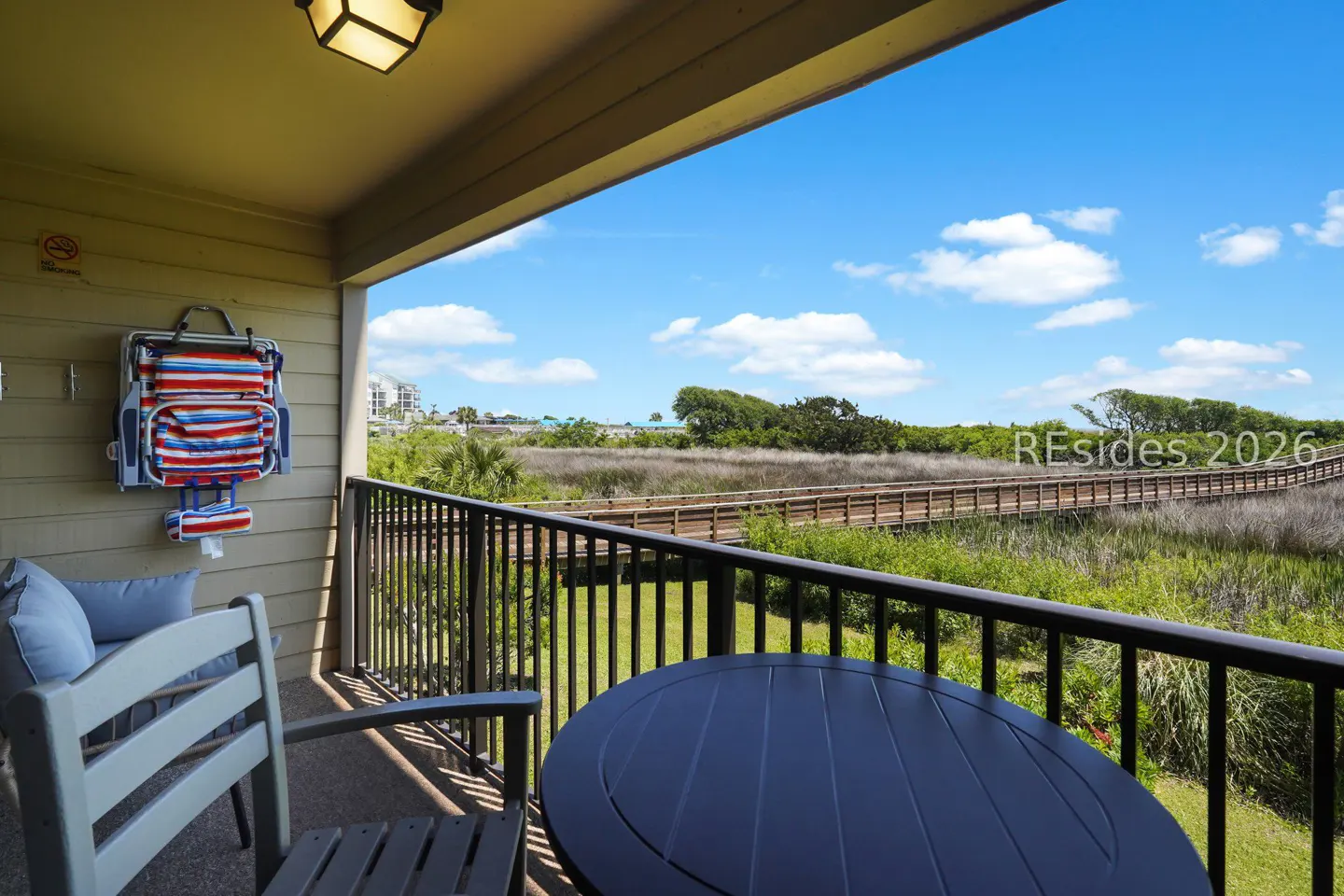 Balcony view with black metal railing, table, and chair. A beach chair hangs on the wall. A boardwalk stretches through green marshland under a blue sky.